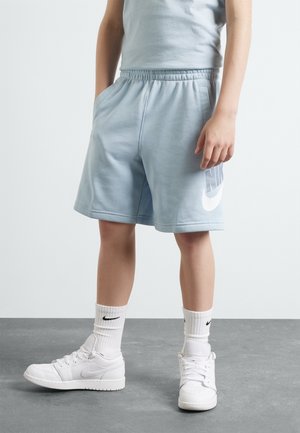 Barn iført lyseblå Nike-shorts, hvide Nike-strømper og hvide Nike-sneakers, stående på en grå gulv.