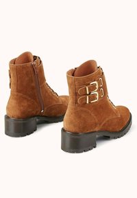Bottines en daim marron avec un bout arrondi, fermeture éclair, lacets à l'avant et deux boucles dorées sur le côté ; talon compas en caoutchouc noir.