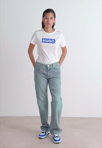 Bílý tričko s modrým logem "HUGO", v kombinaci se světle modrými vzorovanými džíny a modro-bílými teniskami. Modelka stojí s rukama na bocích.