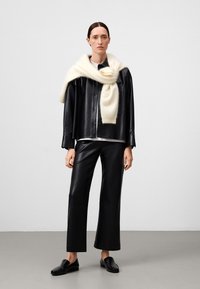 Camicia nera in pelle con bottoni, pantaloni neri in pelle a vestibilità larga, sciarpa di lana bianca drappeggiata sulle spalle, scarpe nere senza lacci, sfondo neutro.