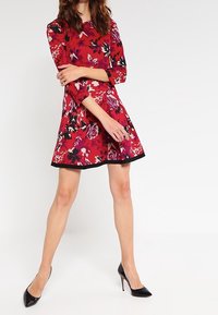 Robe fleurie rouge avec manches trois quarts, jupe évasée, ourlet noir, et un mélange de motifs floraux blancs, roses et bleu marine. Portée avec des escarpins noirs.