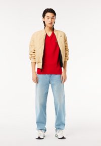 Beige quiltet bomberjakke, rød V-hals t-shirt, lys blå lige ben jeans, hvide sneakers med accenter. Model står foran en hvid baggrund.
