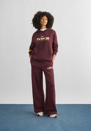 Frau steht mit den Händen in den Taschen, trägt einen weinroten Nike-Hoodie und passende weit geschnittene Hosen, weiße Sneaker, vor schlichtem Hintergrund.