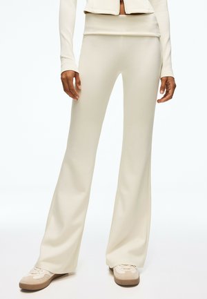 Personne portant un pantalon beige à jambes larges et des baskets assorties, debout devant un fond blanc uni.