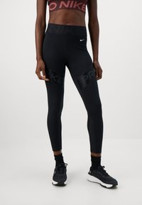 Sportliche Person in schwarzen Nike Pro Leggings, schwarzen Socken, schwarz-weißen Nike Turnschuhen und einem kurzen Nike-Oberteil, steht vor einem weißen Hintergrund.