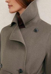 Sivý vlnený kabát s vysokým golierom, ktorý má čierne gombíky a hladkú textúru. Golier je preložený a vrstvený pod tmavým rolákom.