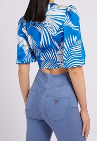Blusa blu cropped con stampa di foglie di palma bianche, abbinata a jeans azzurri a vita alta con un logo triangolare sulla tasca posteriore.