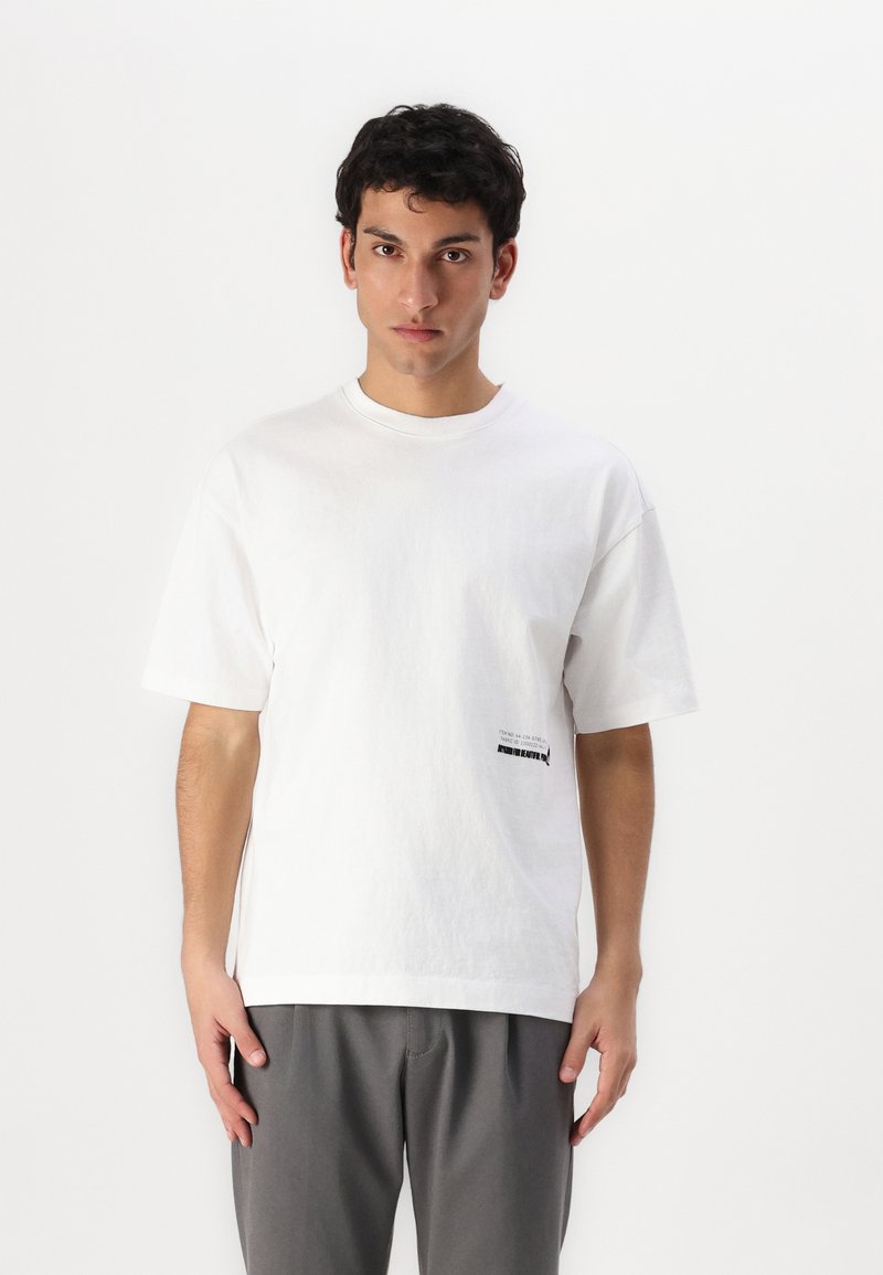 Tricou alb din bumbac, cu guler rotund, mâneci scurte și o mică imprimare neagră în partea de jos a frontalului. Purtați-l cu pantaloni gri.