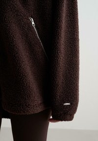 Bruine fleecejacket met een gestructureerd oppervlak, voorzien van een zijrits en een klein zilverkleurig logo-detail op de mouw. Zacht en warm uiterlijk.