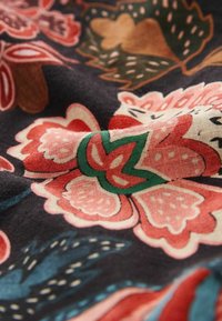 Tissu avec un fond sombre orné de grands motifs floraux en rouge, rose, vert et marron. La texture semble douce et légèrement froissée.