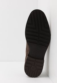 Semelle d'une chaussure marron avec des motifs géométriques, affichant la marque "ecco", positionnée sur des surfaces blanches.