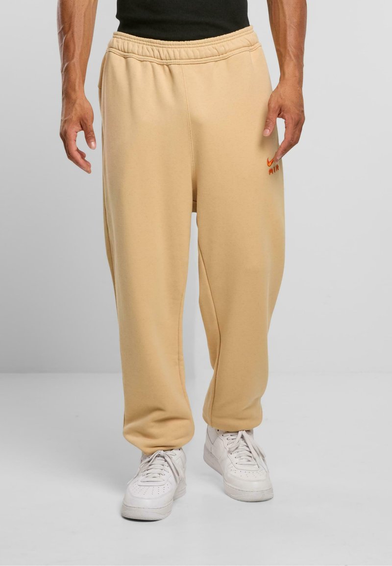 Bežid sweatpants pehmeko kangast, millel on elastne vöökoht, külje taskud ja väikene oranž Nike Air logo vasakul reiel.