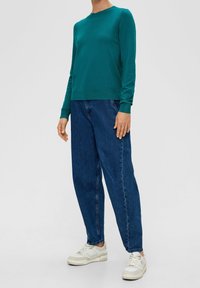 Camisa de manga larga color teal con cuello redondo, combinada con jeans de corte suelto en azul oscuro y zapatillas blancas. Tejido suave, diseño sencillo.