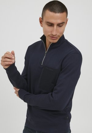 Marinblå sweatshirt med halvmatch, långa ärmar, texturerad tyg och en bröstficka som har en kontrasterande mörk fläck.