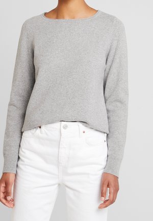 Vrouw draagt een lichtgrijze, gebreide trui met lange mouwen en geribbelde boorden, gecombineerd met witte jeans met een hoge taille.