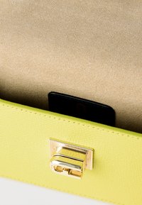 Κίτρινη υφασμάτινη τσάντα με χρυσό ορθογώνιο κλείσιμο τύπου turn-lock σε μπεζ υφασμάτινο φόντο.