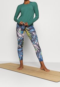 Grön långärmad topp som kombineras med figurnära leggings med ett färgglatt tropiskt bladmönster på en mörk bakgrund.