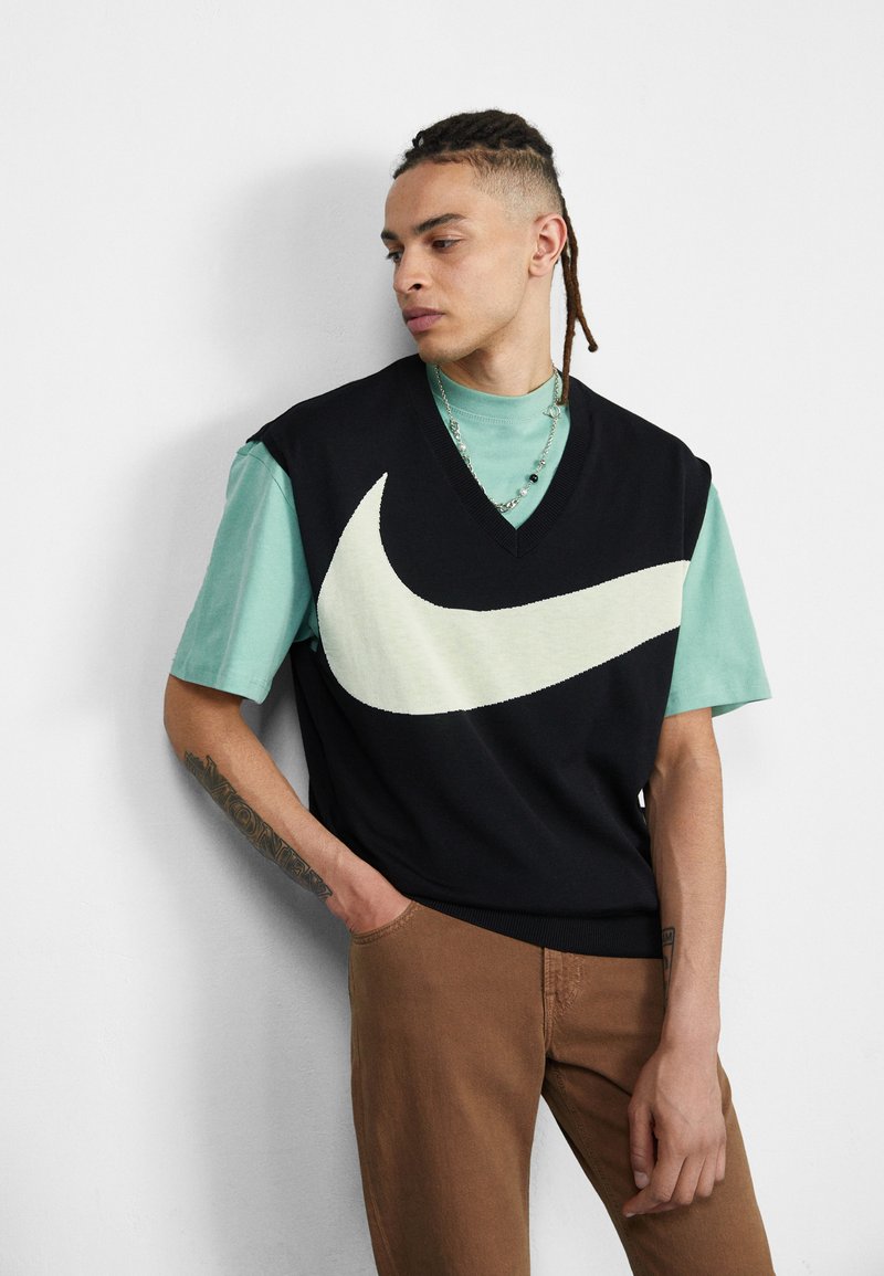 Schwarzer ärmelloser Strickpullover mit großem cremefarbenem Nike-Logo, übereinander getragen mit einem mintgrünen T-Shirt. Braune Hose mit subtiler Textur und geometrischem V-Ausschnitt-Design.
