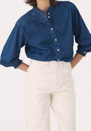 Femme portant un chemisier en denim bleu foncé à boutons, avec un col et des poignets à volants, associé à un pantalon taille haute écru.