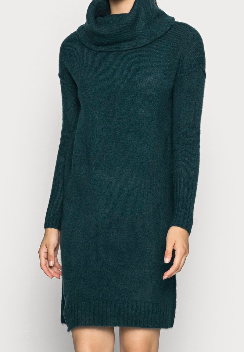 Robe pull en maille vert foncé à manches longues, col roulé ample et poignets et ourlet côtelés, portée par une personne avec les mains relâchées le long du corps.
