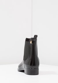 Bottines noires imperméables avec un matériau brillant, un panneau latéral élastique et un petit accent doré. Semelle plate et design à bout arrondi lisse.