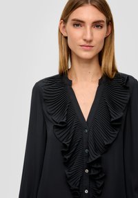 Schwarze Bluse mit einem auffälligen gefältelten Volant an der Vorderseite, V-Ausschnitt und Knopfverschluss mit dekorativen grünen Knöpfen.