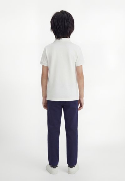 Παιδί με μαύρα μαλλιά στέκεται με την πλάτη γυρισμένη, φορώντας λευκό κοντομάνικο πουκάμισο, μπλε παντελόνι και λευκά αθλητικά παπούτσια μπροστά σε ένα απλό φόντο.