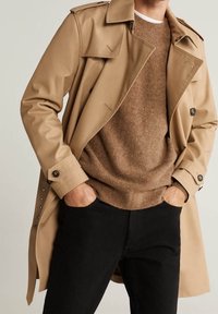 Beiger Trenchcoat mit knöpfbarem Kragen, über einem braunen strukturierten Pullover getragen, kombiniert mit schwarzen Jeans, die einen taillierten Schnitt und klare Linien aufweisen.