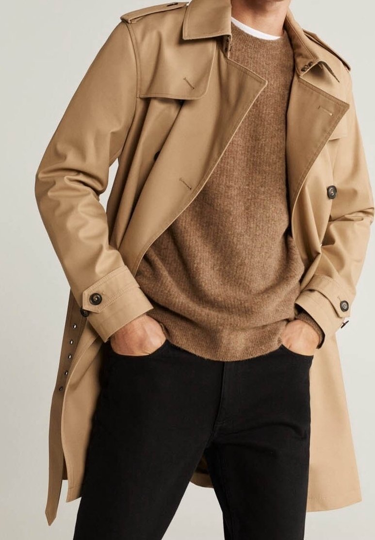 Beiger Trenchcoat mit knöpfbarem Kragen, über einem braunen strukturierten Pullover getragen, kombiniert mit schwarzen Jeans, die einen taillierten Schnitt und klare Linien aufweisen.