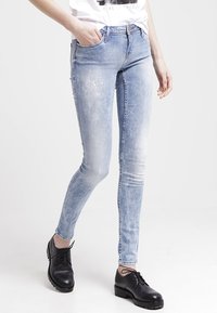 Jeans ajustados de color azul deslavado con un sutil detalle de salpicaduras de pintura, combinados con zapatos negros de cordones y una camiseta gráfica blanca metida por dentro.
