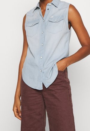 Modrá světlá košile bez rukávů s knoflíky a kapsami na hrudi, nošená s volnými hnědými kalhotami s vysokým pasem, jedna ruka v kapse.