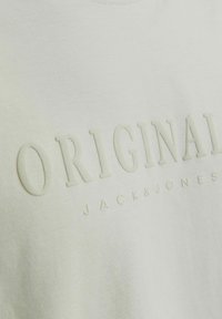 Gaiza zaļā kokvilnas t-kreklu, kuram ir izvirzīts "ORIGINAL" teksts un "JACK & JONES" logo maigi atbilstošā krāsā. Gluda tekstūra.