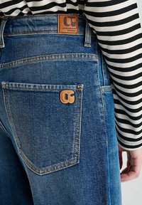 Jean en denim bleu avec une étiquette en cuir visible, présentant des coutures orange et une poche arrière avec un logo distinctif, porté avec une chemise rayée.