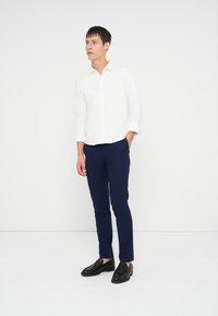 Giovane uomo in piedi con una mano in tasca, indossando una camicia bianca a maniche lunghe, pantaloni blu navy e mocassini neri contro uno sfondo bianco.