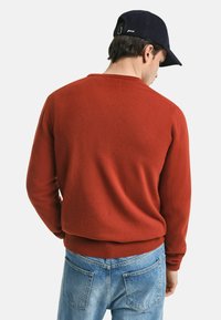 Roter Strickpullover mit rundem Halsausschnitt und gerippten Bündchen, kombiniert mit hellblauen Jeans. Der Stoff wirkt weich und strukturiert.