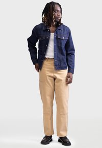 Denimjakke med mørk blå stoff, to brystlommer og kobbersting, parret med beige bukser med store sidelommer. Svarte sko.