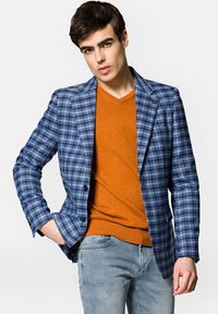Niebiesko-biała marynarka w kratę z dwoma guzikami, noszona na pomarańczowym swetrze. Model ma również na sobie jasnoniebieskie dżinsy. Czyste linie i dobrze zbudowany krój.