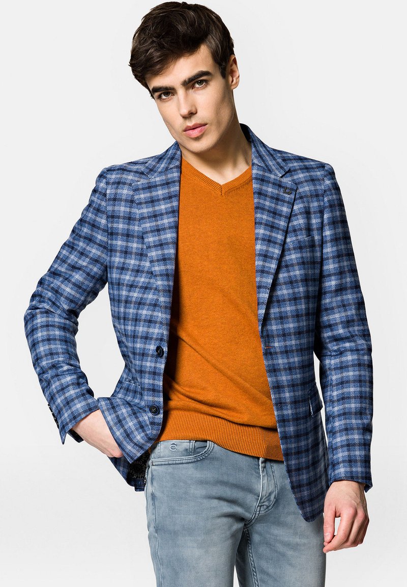 Niebiesko-biała marynarka w kratę z dwoma guzikami, noszona na pomarańczowym swetrze. Model ma również na sobie jasnoniebieskie dżinsy. Czyste linie i dobrze zbudowany krój.