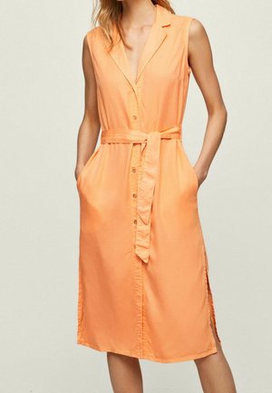 Sukienka bez rękawów w koralowo-pomarańczowym kolorze, zapinana na guziki, z kołnierzem, paskiem do zawiązania w talii, bocznymi rozcięciami i kieszeniami, noszona przez stojącą osobę.