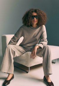 Béžový pletený sveter s textúrou a zodpovedajúce široké nohavice s rebrovaným golierom, nosený s tmavohnedými plochými topánkami, sediaci na svetlej, hebkej stoličke.