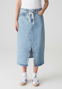 Gonna midi in denim azzurro chiaro, con spacco frontale, vita regolabile con cordoncino e tasche tradizionali. Abbinata a sneakers bianche e calzini.