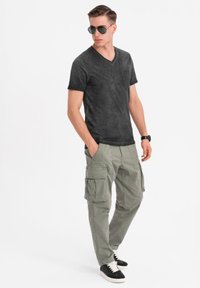 Černé tričko s výstřihem do V a kapsou, olivové cargo kalhoty se bočními kapsami a černé tenisky s bílou podrážkou; model nosí sluneční brýle.