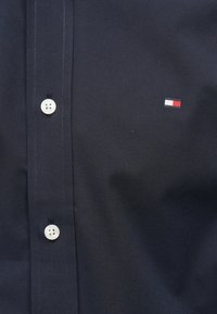 Chemise boutonnée bleu marine foncé en tissu lisse, avec des boutons blancs et un petit logo brodé avec des accents rouges, blancs et bleus.