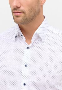 Witte blouse met kleine zwarte stippen, voorzien van een button-down kraag en blauwe knopen. Glad van textuur met een getailleerde pasvorm.