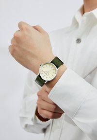 Silver watch with a cream face, green fabric strap, and black markings, worn on a wrist with a white long-sleeve shirt.