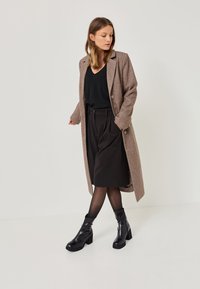 Rjav volnen mešan coat z izrezanim ovratnikom, črn zgornji del, nabran črn krilo in črne gležnjarje. Gladka tekstura, krojen fit.