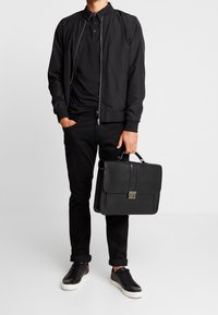 Hombre con chaqueta bomber negra, polo negro, pantalones negros arremangados, zapatillas negras, sosteniendo un maletín de cuero negro con cierre de metal.