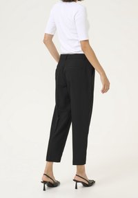 Pantalon noir sur mesure avec jambes fuselées, deux poches arrière et une texture lisse, associé à un haut blanc ajusté et des sandales à talons noires.