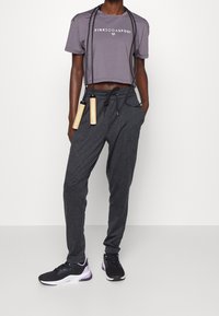 Person som bär mörkgrå joggers, en kort lila T-shirt med texten "PINK SODA SPORT", svarta sneakers, håller ett svart hopprep med trähandtag.