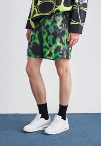 Homme portant un short Nike Air vert et noir à motifs, des chaussettes noires, des baskets blanches Nike et un t-shirt noir et jaune à manches longues, debout sur un tapis bleu.